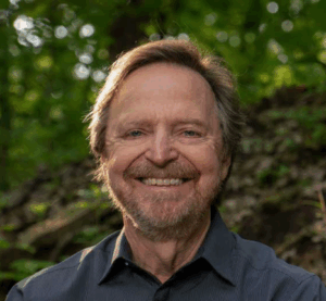 A middle-aged man with light brown hair and a beard smiles outdoors, wearing a dark shirt. The background is lush and green with trees and foliage, suggesting a natural, wooded setting.